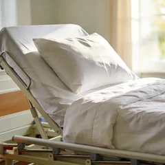 An adjustable home hospital bed with the head section raised, made up with white linens in a bright, welcoming home bedroom